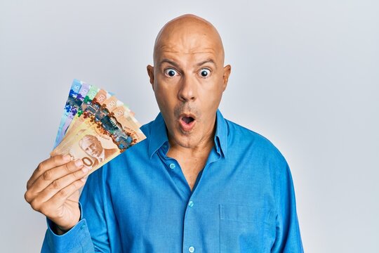 Middle age bald man holding canadian dollars scared and amazed with open mouth for surprise, disbelief face