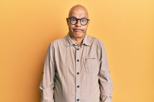 Middle age bald man wearing casual clothes and glasses puffing cheeks with funny face. mouth inflated with air, crazy expression.