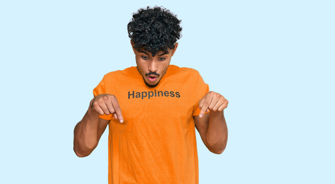Young arab man wearing tshirt with happiness word message pointing down with fingers showing advertisement, surprised face and open mouth