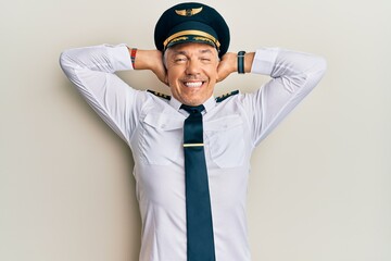 Handsome middle age mature man wearing airplane pilot uniform relaxing and stretching, arms and hands behind head and neck smiling happy