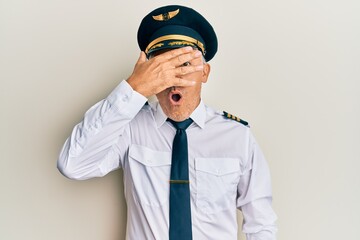 Handsome middle age mature man wearing airplane pilot uniform peeking in shock covering face and eyes with hand, looking through fingers with embarrassed expression.