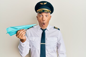 Handsome middle age mature man wearing airplane pilot uniform holding paper plane scared and amazed...