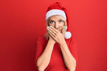 Young blonde girl wearing christmas hat shocked covering mouth with hands for mistake. secret concept.