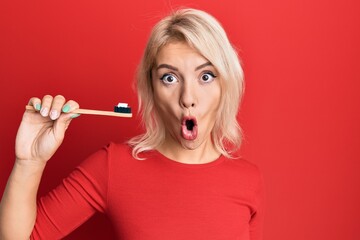 Young blonde girl holding toothbrush with toothpaste scared and amazed with open mouth for surprise, disbelief face