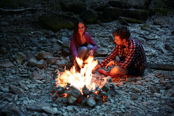 Young Caucasian man and woman sitting near a bonfire in the evening in the summer mountains.