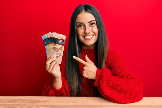 Beautiful hispanic woman holding canadian dollars smiling happy pointing with hand and finger