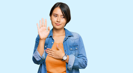 Young beautiful hispanic woman with short hair wearing casual denim jacket swearing with hand on...