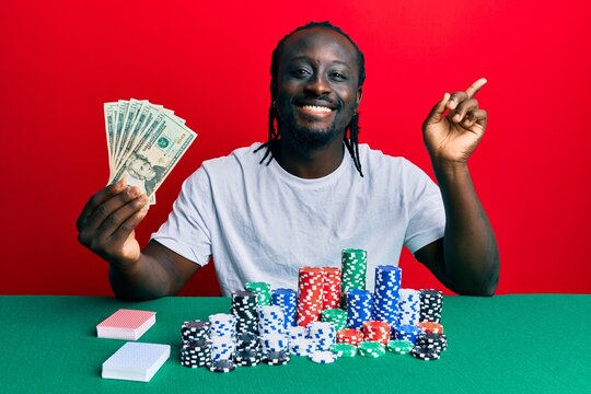 Handsome young black man playing poker holding 20 dollars banknotes smiling happy pointing with hand and finger to the side