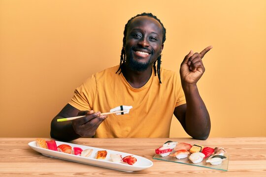Handsome young black man eating sushi sitting on the table smiling happy pointing with hand and finger to the side
