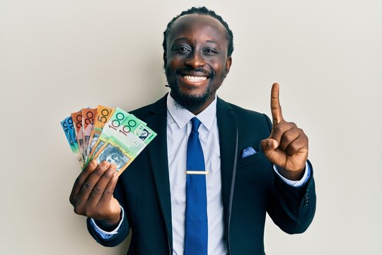 Handsome young black man wearing business suit and tie holding australian dollars smiling with an idea or question pointing finger with happy face, number one
