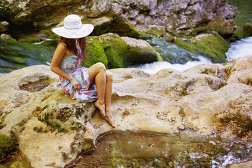 Young caucasian woman in a dress and hat resting on stones near a mountain river.