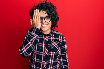 Young hispanic woman with curly hair wearing casual clothes and glasses covering one eye with hand, confident smile on face and surprise emotion.