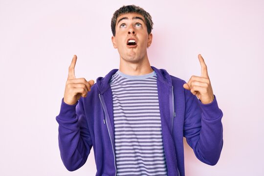 Young handsome man wearing casual purple sweatshirt amazed and surprised looking up and pointing with fingers and raised arms.