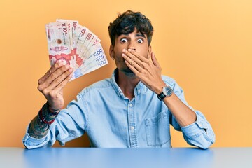 Young hispanic man holding colombian pesos sitting on the table covering mouth with hand, shocked and afraid for mistake. surprised expression