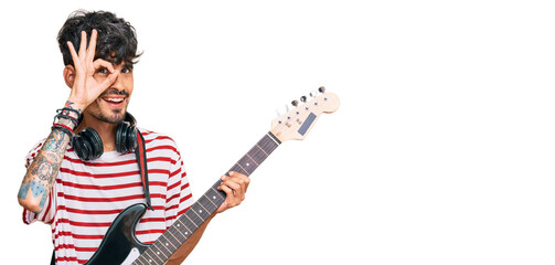 Fototapeta premium Young hispanic man playing electric guitar smiling happy doing ok sign with hand on eye looking through fingers