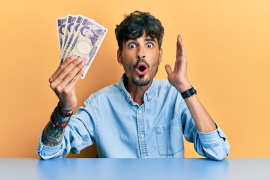 Young hispanic man holding japanese yen banknotes sitting on the table scared and amazed with open mouth for surprise, disbelief face