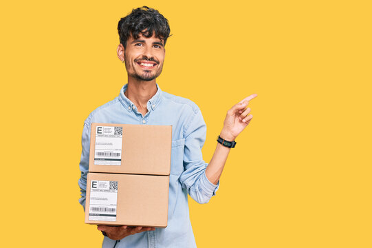 Young hispanic man holding delivery package smiling happy pointing with hand and finger to the side