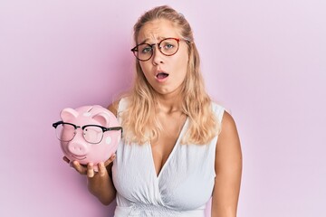 Young blonde girl holding piggy bank with glasses scared and amazed with open mouth for surprise, disbelief face