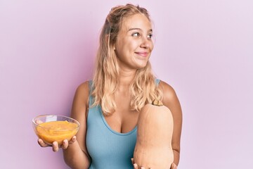 Young blonde girl holding fresh pumpkin and soup smiling looking to the side and staring away thinking.