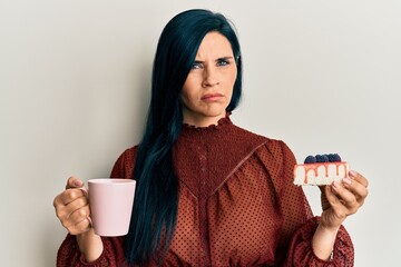 Young caucasian woman holding cup of coffee and cake slice skeptic and nervous, frowning upset...