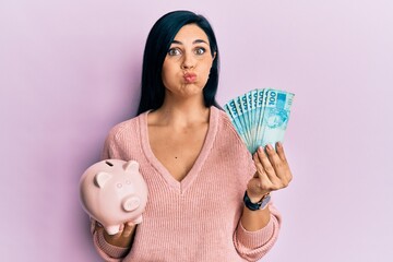 Young caucasian woman holding brazil real banknotes and piggy bank puffing cheeks with funny face. mouth inflated with air, catching air.