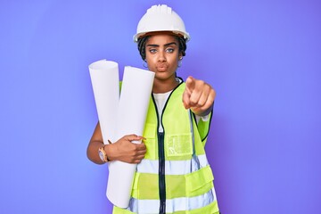 Young african american woman with braids wearing safety helmet holding blueprints pointing with finger to the camera and to you, confident gesture looking serious