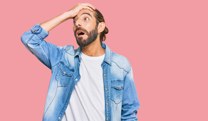 Attractive man with long hair and beard wearing casual denim jacket surprised with hand on head for mistake, remember error. forgot, bad memory concept.