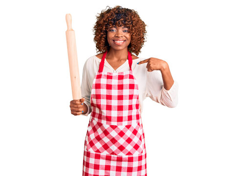 Young african american woman wearing apron holding rolling pin pointing finger to one self smiling happy and proud