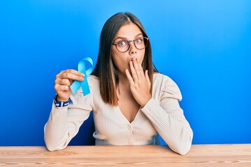 Young caucasian woman holding blue ribbon covering mouth with hand, shocked and afraid for mistake....
