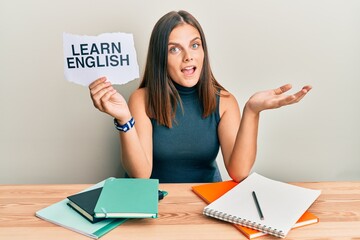 Young caucasian woman holding learn english paper while studying celebrating achievement with happy smile and winner expression with raised hand