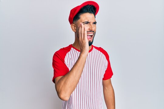 Young man with beard wearing baseball uniform shouting and screaming loud to side with hand on mouth. communication concept.
