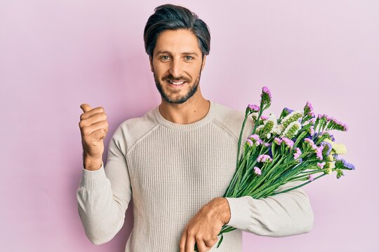 Young hispanic man holding flowers pointing thumb up to the side smiling happy with open mouth