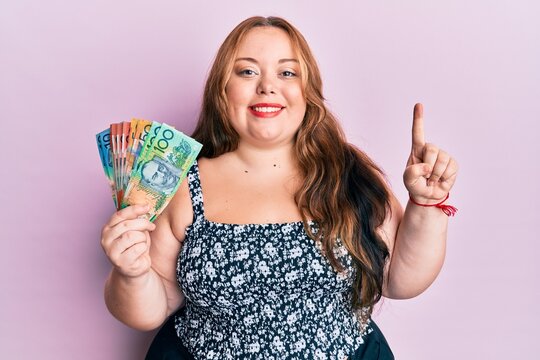 Plus size caucasian young woman holding australian dollars smiling with an idea or question pointing finger with happy face, number one