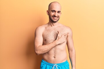 Young handsome bald man wearing swimwear shirtless smiling cheerful pointing with hand and finger up to the side