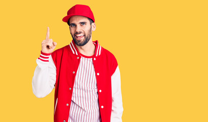 Young handsome man with beard wearing baseball jacket and cap pointing finger up with successful idea. exited and happy. number one.