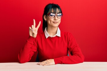 Young brunette woman with bangs wearing glasses sitting on the table showing and pointing up with fingers number two while smiling confident and happy.