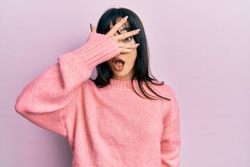 Young brunette woman with bangs wearing casual winter sweater peeking in shock covering face and eyes with hand, looking through fingers with embarrassed expression.