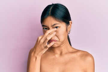 Young chinese woman standing topless showing skin smelling something stinky and disgusting, intolerable smell, holding breath with fingers on nose. bad smell