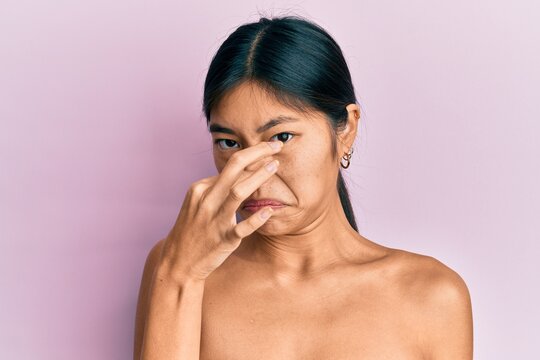 Young Chinese Woman Standing Topless Showing Skin Smelling Something Stinky And Disgusting, Intolerable Smell, Holding Breath With Fingers On Nose. Bad Smell