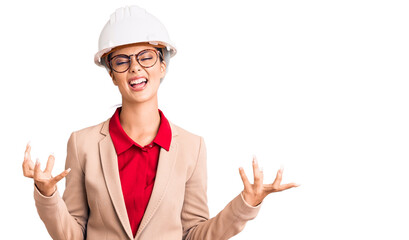 Young beautiful woman wearing architect hardhat crazy and mad shouting and yelling with aggressive expression and arms raised. frustration concept.