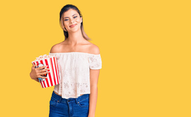 Young beautiful woman holding popcorn looking positive and happy standing and smiling with a confident smile showing teeth
