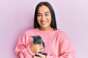 Young hispanic girl smiling happy holding cute cat over isolated pink background.