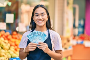 Young latin shopkeeper girl smiling happy holding brazil real banknotes at the fruit store. - obrazy, fototapety, plakaty