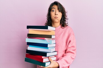Young hispanic woman holding a pile of books puffing cheeks with funny face. mouth inflated with air, catching air.