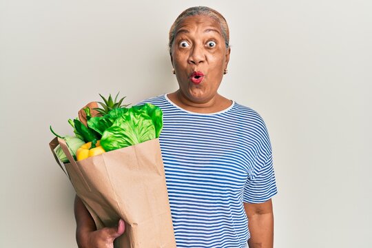 Senior African American Woman Holding Paper Bag With Bread And Groceries Scared And Amazed With Open Mouth For Surprise, Disbelief Face