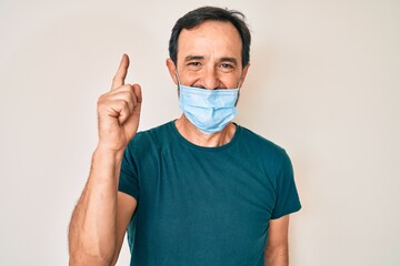 Middle age hispanic man wearing medical mask smiling with an idea or question pointing finger with happy face, number one