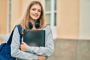 Beautiful caucasian student teenager using headphones holding binder at the city.