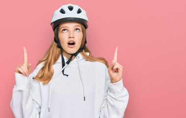 Beautiful young caucasian girl wearing bike helmet amazed and surprised looking up and pointing with fingers and raised arms.
