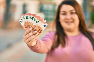 Young hispanic plus size woman smiling happy holding 10 euro banknotes at the city.
