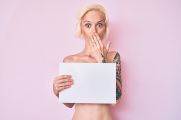 Young blonde woman with tattoo standing shirtless holding empty blank banner covering mouth with...